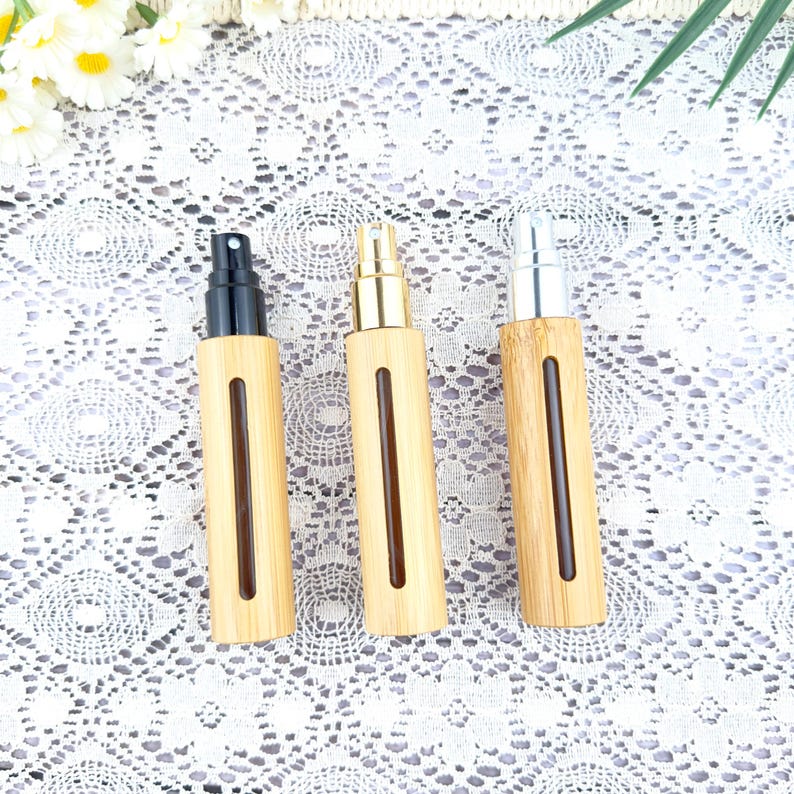 May include: Three bamboo spray bottles with different colored tops: black, gold, and silver. Each bottle has a rectangular window showing the liquid level. The bottles are arranged on a white lace surface.