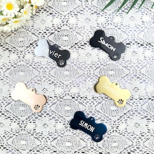 Puede incluir: Etiquetas para mascotas en forma de hueso en varios colores: negro, dorado, azul marino y oro rosa. Cada etiqueta tiene un nombre grabado, como "Simon" o "Xavier", y una huella de pata. Las etiquetas son para mascotas.