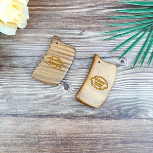May include: Two wooden, light brown Gua Sha tools with engraved text. One reads "Anthony" with a heart design, the other reads "Johnny Best Man Wedding". Each has a small hole at the top. The tools are on a wooden surface.