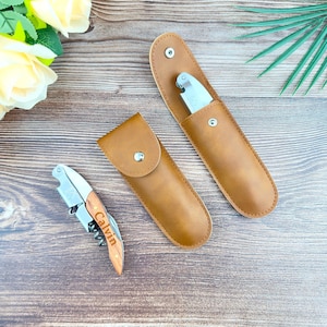 Puede incluir: Un sacacorchos de acero inoxidable con mango de madera, grabado con el nombre "Calvin", junto a dos estuches de cuero marrón, uno abierto y otro cerrado. Los estuches tienen cierre de botón a presión.