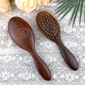 May include: Two dark brown wooden hairbrushes. One has the engraved text "Johnny Best Man 07.11.2004". The other has wooden bristles. Both brushes are oval-shaped and suitable for hair care.