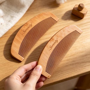May include: Two curved wooden combs with a light brown finish and a smooth texture. Each comb features the word Serenity engraved in a dark script font on the upper surface. The combs have fine, evenly spaced teeth along the bottom edge.