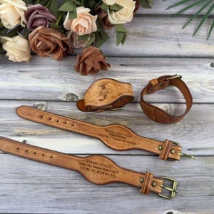 Op de afbeelding: Een verzameling gepersonaliseerde lederen accessoires, waaronder twee riemen, een manchet en een armband. De items zijn lichtbruin van kleur en voorzien van gegraveerde tekst en metalen gespen. Op de achtergrond staat een boeket rozen.
