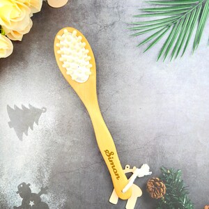 May include: A light-colored wooden massage brush with a long handle and an oval head covered in small, white, rounded massage nodes. The name "Simon" is engraved on the handle. A small wooden reindeer ornament hangs from the handle.