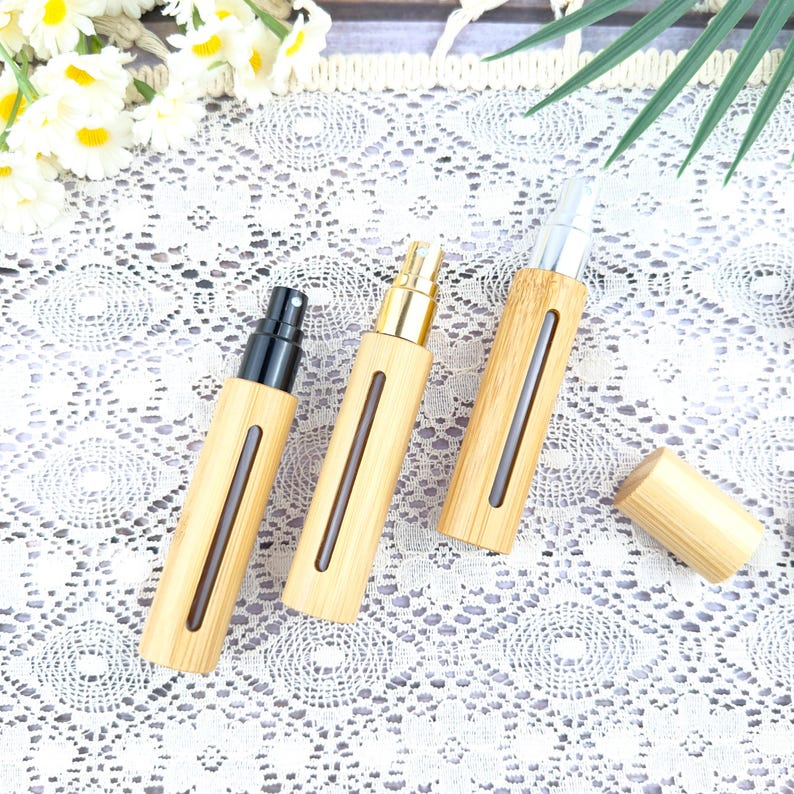 May include: Three bamboo-style perfume atomizers with different colored spray tops: black, gold, and silver. Each atomizer has a clear window and a matching bamboo cap. The atomizers are arranged on a white lace surface.
