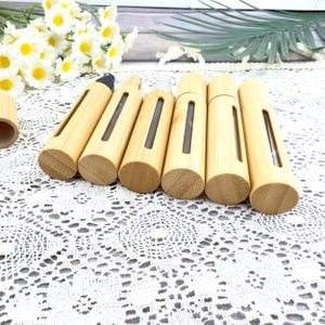 May include: A collection of bamboo essential oil roller bottles. Each bottle has a natural wood finish with a rectangular window to view the contents. The bottles are cylindrical with a rounded base and a cap. The background includes white flowers and greenery.