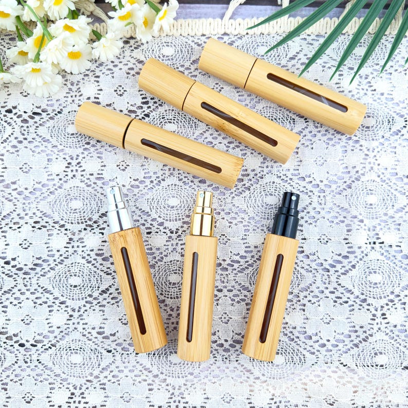 May include: A collection of bamboo essential oil spray bottles. The bottles have a natural wood finish with a rectangular window and a silver, gold, or black spray nozzle. The bottles are arranged on a white lace surface with flowers and greenery.