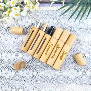 May include: A collection of bamboo perfume atomizers with silver, gold, and black spray tops. The atomizers are engraved with the names "Simon" and "Juan" in a gold script. The atomizers are displayed on a white lace surface.