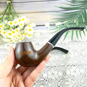 Puede incluir: Una pipa de madera marrón con el nombre "SIMON" grabado en la cazoleta. La pipa tiene una boquilla negra y una banda plateada. La pipa está sujeta en una mano.