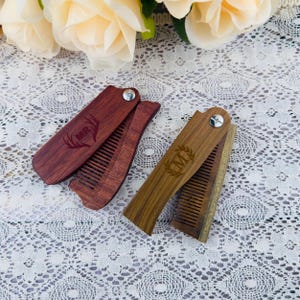 May include: Two wooden folding combs, one in a dark reddish-brown and the other in a light brown. The dark comb has the initials "MR" with a deer antler design, while the light comb has the initial "M" within a laurel wreath.