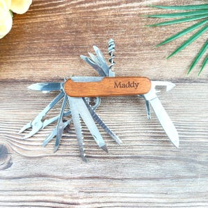 May include: A multi-tool with a wooden handle and various stainless steel implements, including a knife, saw, and bottle opener. The name "Maddy" is engraved on the handle. The tool is displayed open, showcasing its many functions.