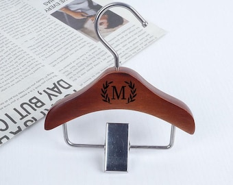 May include: A wooden clothes hanger with a metal hook and clip. The hanger is a dark brown color and features a laurel wreath design with the letter "M" in the center. The hanger is designed to hold clothing.