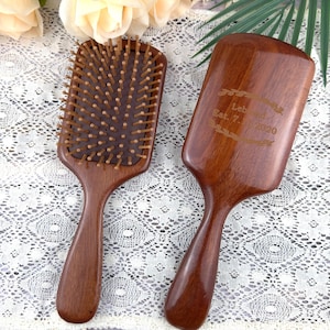 May include: Two wooden hairbrushes. One has wooden bristles, the other has the text "Leband Est. 7. 2020" engraved on the surface. Both brushes are dark brown with ergonomic handles. The brushes are designed for hair care.