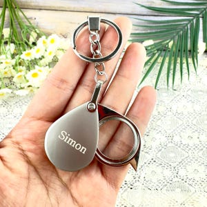Może przedstawiać: Srebrny brelok do kluczy z lupą i zawieszką w kształcie łzy z wygrawerowanym imieniem "Simon". Brelok ma dzielony pierścień i mały łańcuszek łączący zawieszkę i lupę.