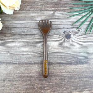May include: A wooden back scratcher with a handle and a claw-like head. The scratcher is a dark brown color and has the word "Remino" printed on the handle in a gold color. The scratcher is on a wooden surface.