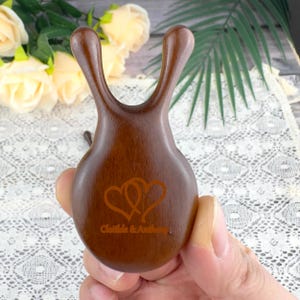 Puede incluir: Una herramienta de masaje de madera marrón oscuro con una forma única, con dos asas curvas y una base redondeada. La base tiene un diseño de corazón y el texto "Clotilde & Anthony".