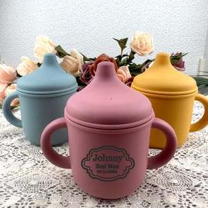 May include: Three pastel-coloured silicone baby sippy cups: blue, pink, and yellow. The pink cup has the text "Johnny Best Man 07.11.2024" in a decorative frame. Each cup has a lid and two handles.