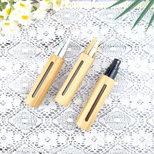 May include: Three bamboo spray bottles with silver, gold, and black spray tops. The bottles have a rectangular shape with a clear window to view the contents. The bottles are arranged on a white lace surface.