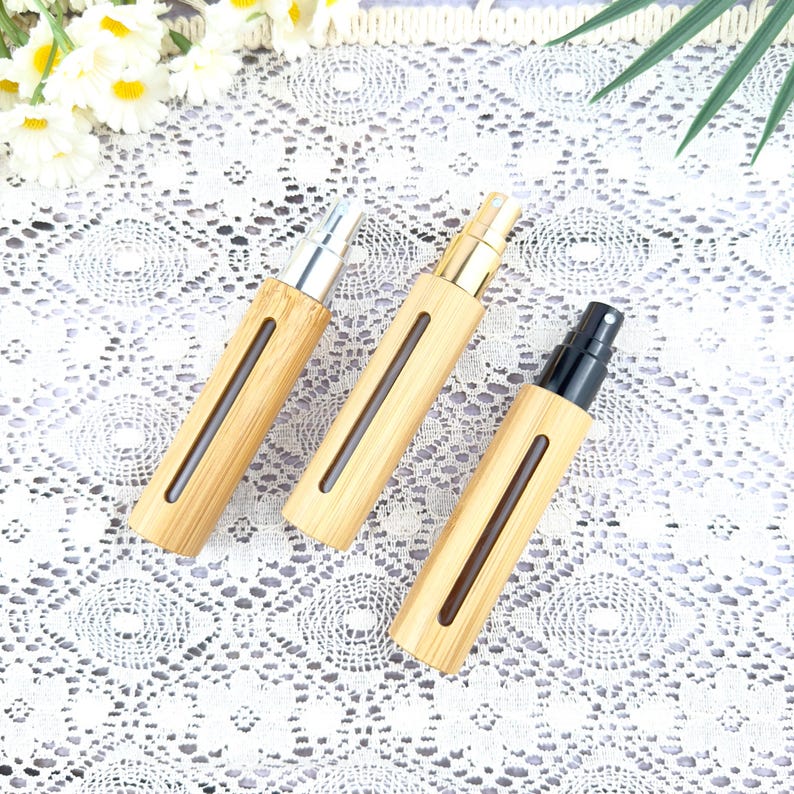 May include: Three bamboo spray bottles with silver, gold, and black spray tops. The bottles have a rectangular shape with a clear window to view the contents. The bottles are arranged on a white lace surface.