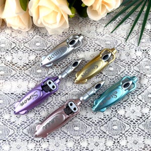 May include: An assortment of metallic bottle openers in silver, gold, purple, rose gold, and teal. Each opener has a unique design with the name "Simon" printed on it. The openers are arranged on a white lace surface.