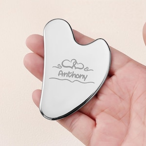 Op de afbeelding: Een zilveren gua sha-tool met een hartontwerp en de naam "Anthony" gegraveerd. De tool wordt in een hand gehouden en is ontworpen voor gezichtsmassage en huidverzorging.