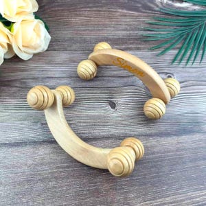 May include: Two wooden massage rollers with curved handles and grooved spheres. One roller has the name "Simon" engraved in yellow. The rollers are made of light-colored wood and are designed for muscle relaxation and therapy.