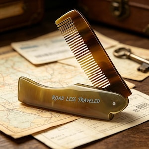 Peut inclure: Un peigne pliant avec une finition lisse, translucide et semblable à de la corne dans des tons ambre et brun. Le manche porte le texte gravé ROAD LESS TRAVELED. Le peigne est présenté en position ouverte, révélant ses dents fines et un pivot.