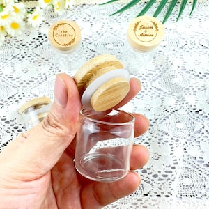 May include: A clear glass jar with a wooden lid is held in a hand. The lid is made of light-colored wood and has a white silicone seal. Two other jars with wooden lids are visible, one with the text "the Creative" and the other with "Jason & Aimee".