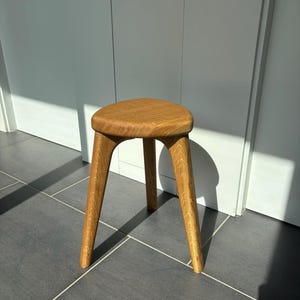 Pode incluir: Um banco de madeira de três pernas com um assento triangular. O banco é feito de madeira clara e tem um acabamento natural. As pernas são anguladas e se abrem para fora. O banco tem aproximadamente 46 cm de altura.