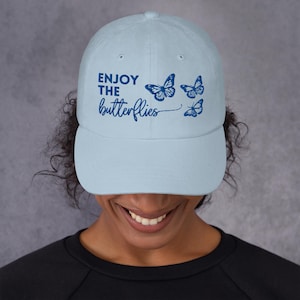Puede incluir: Gorra de béisbol azul claro con el texto "ENJOY THE butterflies" en azul oscuro. La gorra presenta tres ilustraciones de mariposas azules oscuras. El sombrero está hecho de un material suave y tiene una visera curva.