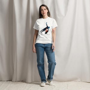 Op de afbeelding: Een wit T-shirt met korte mouwen en een afbeelding van twee figuren in een dans-liftpose. Onder de afbeelding staat de tekst "having the time of my life" in een klein, donker lettertype. Gecombineerd met een blauwe spijkerbroek en witte sneakers.