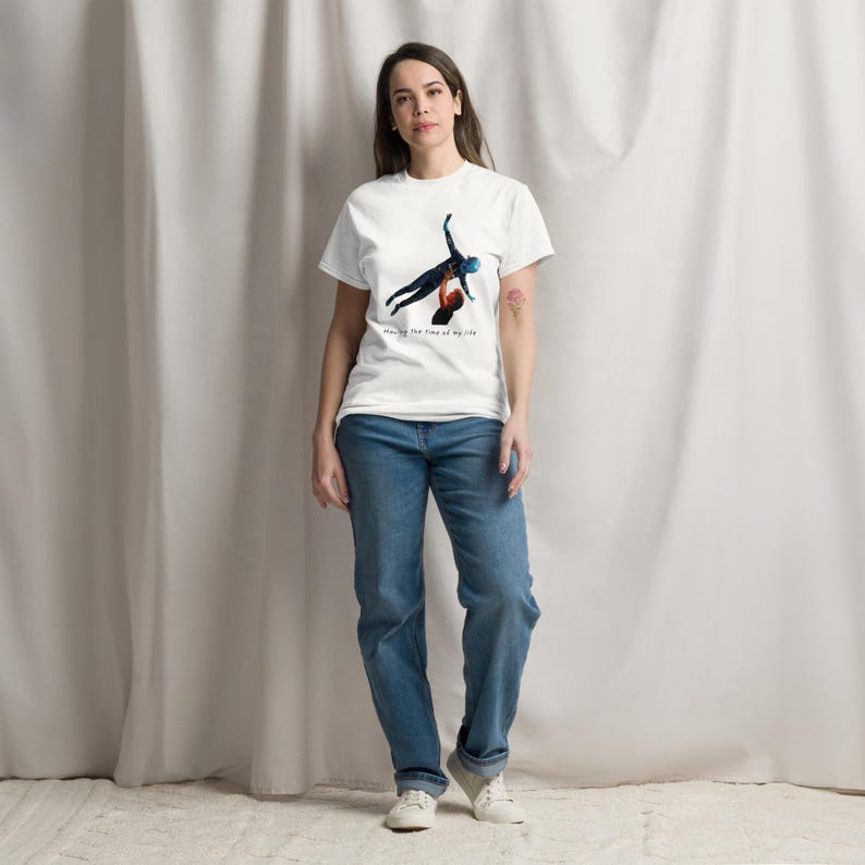 Op de afbeelding: Een wit T-shirt met korte mouwen en een afbeelding van twee figuren in een dans-liftpose. Onder de afbeelding staat de tekst "having the time of my life" in een klein, donker lettertype. Gecombineerd met een blauwe spijkerbroek en witte sneakers.