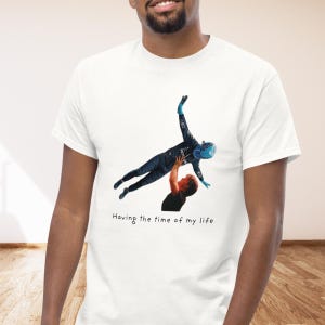 Op de afbeelding: Een wit t-shirt met korte mouwen en een grafische print van een persoon in een donkerblauw racepak en blauwe helm die in de lucht wordt getild door een ander persoon in een zwart shirt. De tekst "Having the time of my life" staat eronder gedrukt.