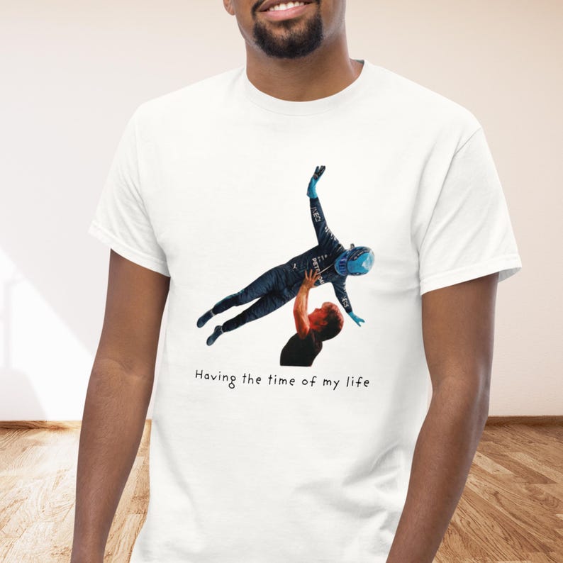 Op de afbeelding: Een wit t-shirt met korte mouwen en een grafische print van een persoon in een donkerblauw racepak en blauwe helm die in de lucht wordt getild door een ander persoon in een zwart shirt. De tekst "Having the time of my life" staat eronder gedrukt.
