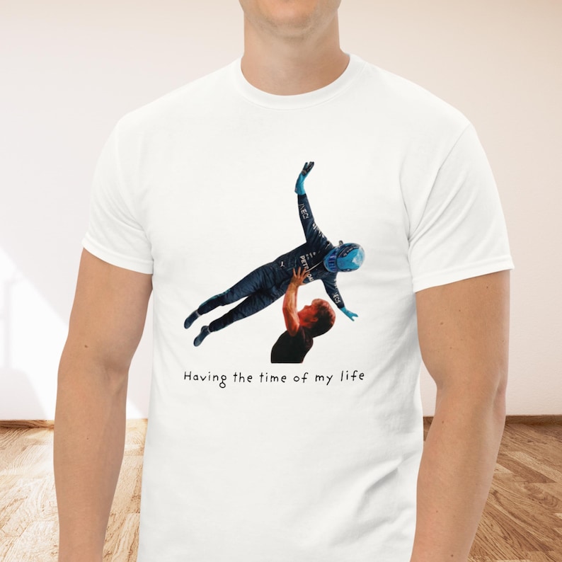 Op de afbeelding: Een wit t-shirt met korte mouwen en een grafische print van een persoon in een donkerblauw racepak die door een ander in de lucht wordt getild. Onder de afbeelding staat de tekst "Having the time of my life" in een eenvoudig zwart lettertype.