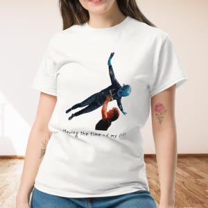 Op de afbeelding: Een wit T-shirt met korte mouwen en een grafische print van een persoon in een donker racepak die door een ander wordt opgetild. De tekst "Having the time of my life" is in het zwart onder de afbeelding gedrukt op het shirt.