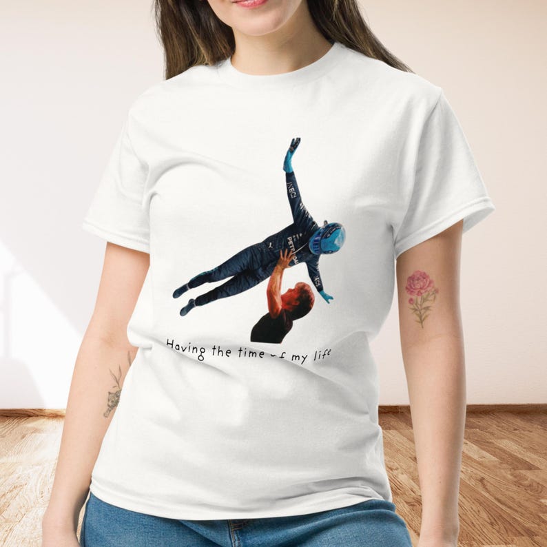 Op de afbeelding: Een wit T-shirt met korte mouwen en een grafische print van een persoon in een donker racepak die door een ander wordt opgetild. De tekst "Having the time of my life" is in het zwart onder de afbeelding gedrukt op het shirt.