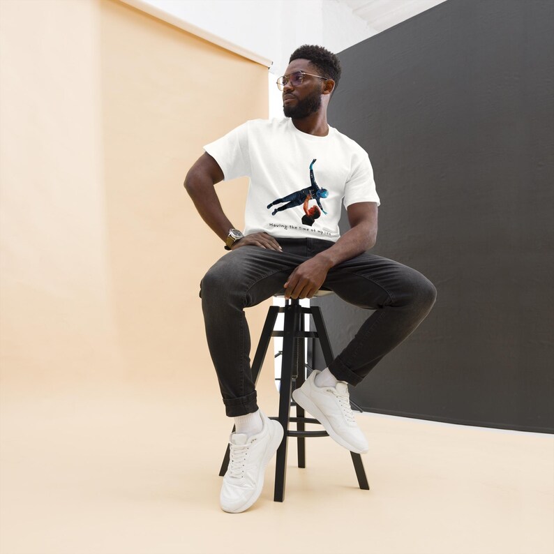 Op de afbeelding: Een wit T-shirt met korte mouwen en een grafische print van twee figuren die een lift uitvoeren, met de tekst "Having the time of my life" onder de illustratie. Het shirt wordt gedragen met een donkergrijze spijkerbroek en witte sneakers.