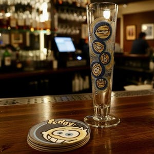 May include: A tall, clear glass with cartoon faces and a matching coaster set on a wooden bar. The glass has a flared base and is decorated with blue and yellow cartoon faces. The coaster has the word "today:" and a smiling cartoon face.