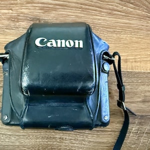 Puede incluir: Funda de cámara Canon de cuero negro con correa para el hombro. El logotipo de Canon en blanco destaca en la parte delantera. La funda es rectangular con bordes redondeados y cierre de solapa.