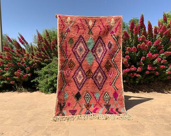 Puede incluir: Una alfombra tejida a mano de color coral con un patrón geométrico de diamantes en morado, turquesa y negro. La alfombra tiene un borde con flecos y se exhibe al aire libre contra un telón de fondo de arbustos en flor y un cielo azul claro.
