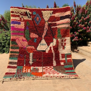 以下が含まれることがあります： 幾何学模様の手織りの鮮やかなラグ。ラグは、白、緑、オレンジ、青のアクセントが付いた深い赤色の背景が特徴です。デザインには、抽象的な形とパターンが含まれており、フリンジエッジが付いています。ラグは屋外に展示されています。