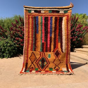 May include: A rectangular, hand-woven rug with a vibrant, geometric design. The rug features vertical stripes in shades of orange, brown, pink, blue, and white. Diamond and rectangular shapes are present in the lower section. The rug is displayed outdoors.