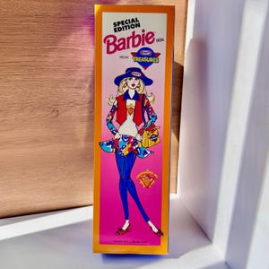 Puede incluir: Una caja de muñeca Barbie vintage con una ilustración vibrante. La caja presenta el texto "SPECIAL EDITION Barbie DOLL FROM TREASURES". La ilustración de Barbie lleva un sombrero azul, un chaleco rojo y pantalones azules. El fondo es rosa y naranja.