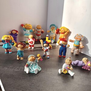 May include: A collection of vintage Cabbage Patch Kids figurines in various outfits and poses. The dolls have different hair colours and wear dresses, overalls, and baseball uniforms. Some dolls are holding toys.