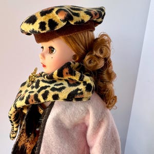 May include: A vintage doll wearing a leopard print beret and scarf, paired with a pink coat. The doll has red hair styled in curls and is adorned with gold-toned earrings and a brooch. The leopard print features black spots on a tan background.