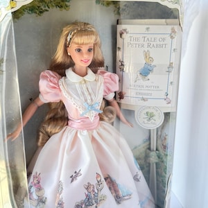 May include: A Barbie doll in a pink and white dress with a Peter Rabbit theme. The doll has blonde hair and is in a clear plastic box. A book titled "The Tale of Peter Rabbit" is visible in the background.