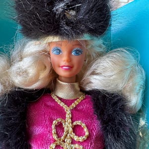 May include: A vintage doll with blonde hair and blue eyes, wearing a pink dress, a black fur coat, and a black hat with a gold band. The doll has a gold necklace and a gold collar.
