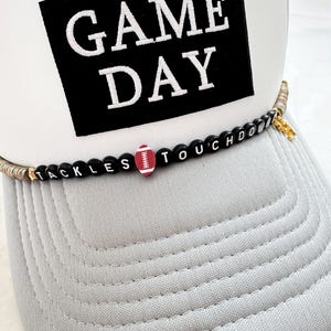 May include: White and gray trucker hat with the text "GAME DAY" in white on a black square. A beaded band spells out "TACKLES TOUCHDOWN" with a football bead. The hat has a curved brim.