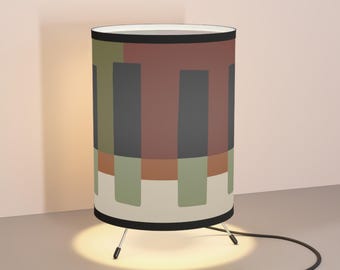 May include: A cylindrical table lamp with a geometric pattern in shades of green, brown, and gray. The lamp has a black trim and three metal legs. A black cord with an on/off switch extends from the base.